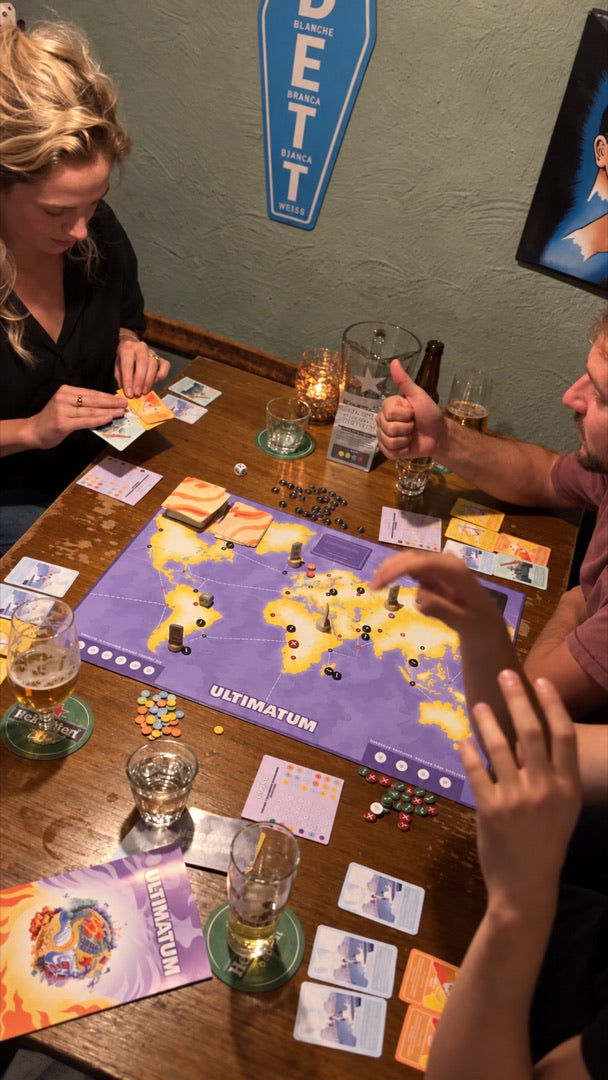 Vier spelers in een café rond het bord van Ultimatum, met bier, een kaarsje en kaarten in de hand